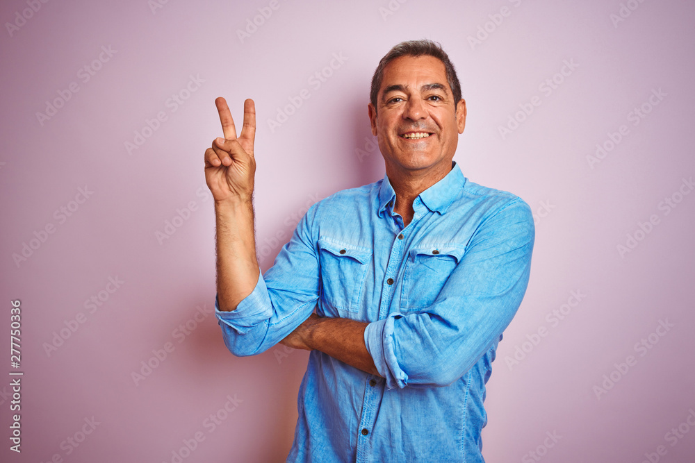 Handsome middle age man wearing blue denim shirt standing over isolated pink background smiling with happy face winking at the camera doing victory sign. Number two.