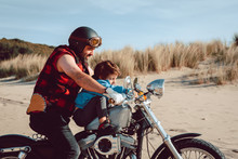 Adult Brutal Father With Son Riding Motorcycle Together On Empty Sandy Beach Of Ocean