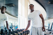 © twinsterphoto - African American man picking up a dumbbell from the rack and looking outside at gym. Male weight training person holding sport equipment with serous look in fitness center