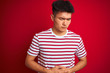 © Krakenimages.com - Young asian chinese man wearing striped t-shirt standing over isolated red background with hand on stomach because nausea, painful disease feeling unwell. Ache concept.
