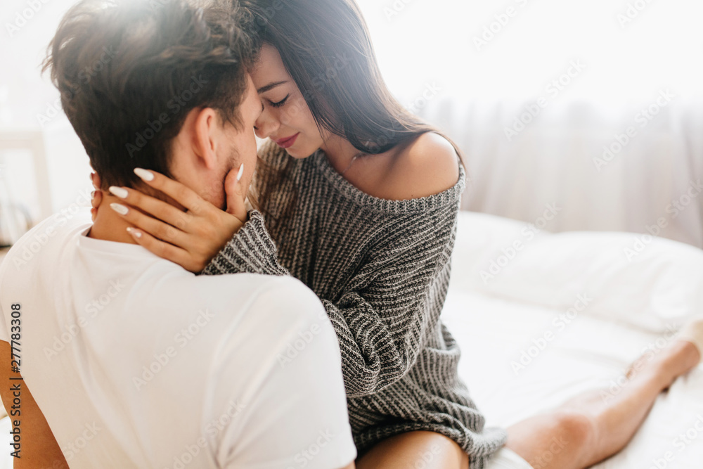 Glamorous tanned girl with white manicure resting with husband in sunday morning. Fascinating european woman holding boyfriend's face with tenderness, sitting on bed. - obrazy, fototapety, plakaty 