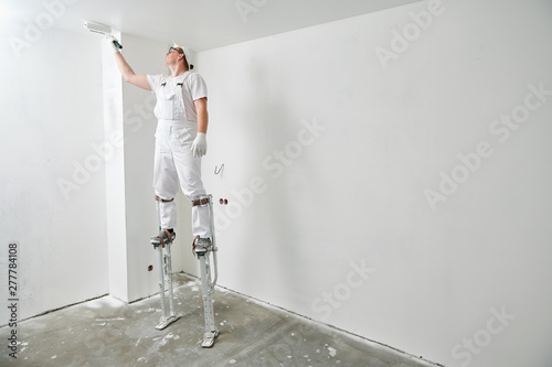 Painter Worker On Stilts With Roller Painting Ceiling Into White