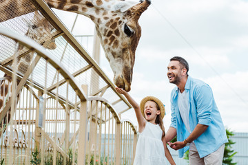 Wall Mural - Selective focus of cheerful man and kid with closed eyes feeding giraffe in zoo