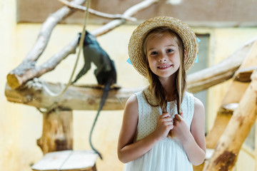 Wall Mural - cheerful kid in straw hat touching hair near monkey in zoo