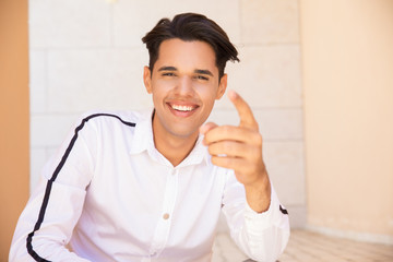 Happy young Latin man pointing at you at wall outdoors. Handsome young guy wearing shirt and looking at camera with wall in background. Promotion concept. Front view.