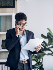 Wall Mural - Asian businessman standing and talking on smart phone contact with client at at modern office