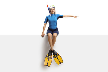 Poster - Young blond female in a wetsuit, a diving mask and flippers sitting on a banner and pointing to the side