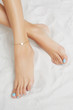 Cropped front shot of girl's legs with pale blue pedicure, wearing golden ankle bracelet, decorated with golden insertion in view of heart. The lady is crossing her legs, lying on the sandy platform.