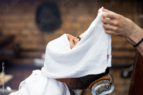 Barber covers the face of a man with a hot towel. Traditional ritual of shaving the beard with hot and cold compresses in a old style barber shop. Hot towel on face before shaving in barber shop