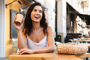 Wall Mural - Portrait of relaxed happy woman with straw bag drinking coffee from paper cup while sitting in cozy cafe