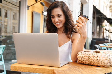 Wall Mural - Portrait of happy seductive woman drinking coffee from paper cup and using laptop while sitting in cozy cafe outdoors