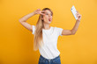 Happy young blonde woman posing isolated over yellow wall background dressed in white casual t-shirt using mobile phone take a selfie.