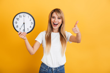 Happy young blonde woman posing isolated over yellow wall background dressed in white casual t-shirt holding clock showing thumbs up.