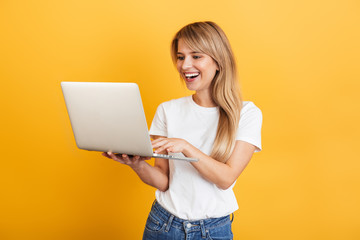 Wall Mural - Positive optimistic emotional young blonde woman posing isolated over yellow wall background dressed in white casual t-shirt using laptop computer.