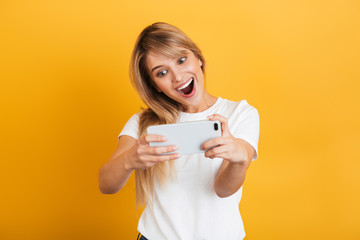 Sticker - Happy emotional young blonde woman posing isolated over yellow wall background dressed in white casual t-shirt using mobile phone play games.