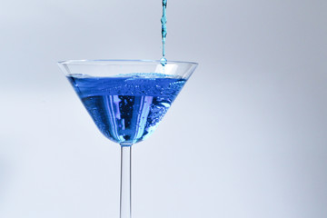 Cocktail with blue liquid in glass. Glass with blue water pouring with liquid with splashes and drops. Martini glass filling with alcohol with splashes on white background. Refreshing drink concept.