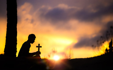 Canvas Print - Silhouette of young male christian sitting and holding a cross for blessing from god with light of sunset background, christian hope concept.