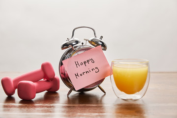 silver alarm clock with happy morning lettering on pink sticky note near pink dumbbells and orange juice on wooden table isolated on grey