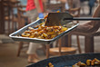 Roast potatoes in a large pan. Delicious food at the festival.A man pours a potato into a metal tray. Blurred people eat at the table in the background.