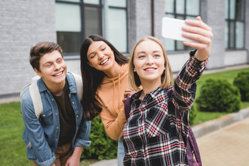 Wall Mural - cheerful teenagers holding smartphone, taking selfie and smiling outside