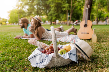 picnic basket in focus, young couple on the grass in the park relaxing with guitar on background