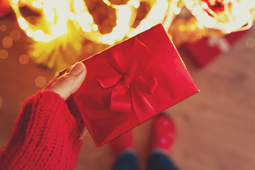 Woman hand holding red Christmas gift box wrapped red ribbon against background of Christmas tree lights. Christmas or New Year celebration concept. Copy space. Selective focus