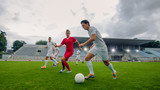 Fototapeta Sport - Professional Soccer Player Leads with a Ball, Masterfully Dribbling and Bypassing Sliding Tackles of His Opponents. Two Professional Football Teams Playing. Low Angle Shot.