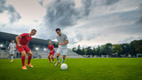 Fototapeta Sport - Professional Soccer Player Leads with a Ball, Masterfully Dribbling and Bypassing Sliding Tackles of His Opponents. Two Professional Football Teams Playing. Low Angle Shot.