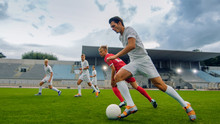 Professional Soccer Player Leads With A Ball, Masterfully Dribbling And Bypassing Sliding Tackles Of His Opponents. Two Professional Football Teams Playing. Low Angle Shot.