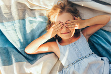 Beautiful Blonde Little Girl Lying On The Blanket, Cover Her Eyes From The Sun With Both Hands, Enjoying Summer Day. Adorable Child Having Fun And Plaiyng Peekaboo Outdoors In The Park