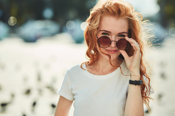 Beautiful red-haired girl having fun on the street. The girls have a beautiful figure, a white T-shirt and jeans with sunglasses