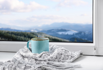 Hot drink and warm scarf near window with view of winter mountain landscape