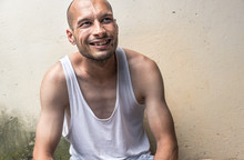 Homeless Man, Young Skinny Anorexic Bald Positive And Happy Smiling Homeless Man Sitting On The Urban Street In The City Or Town Near White Wall With Big Smile, Homelessness Social Documentary Concept