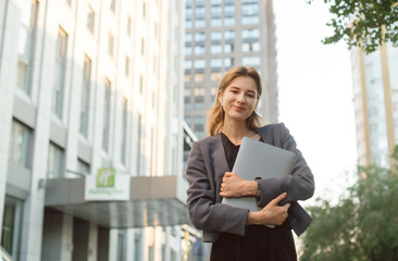 Smiling businesswoman carry laptop using wireless headphones outdoor modern city urban life concept. Positive female office worker in front of the skyscraper and office building.