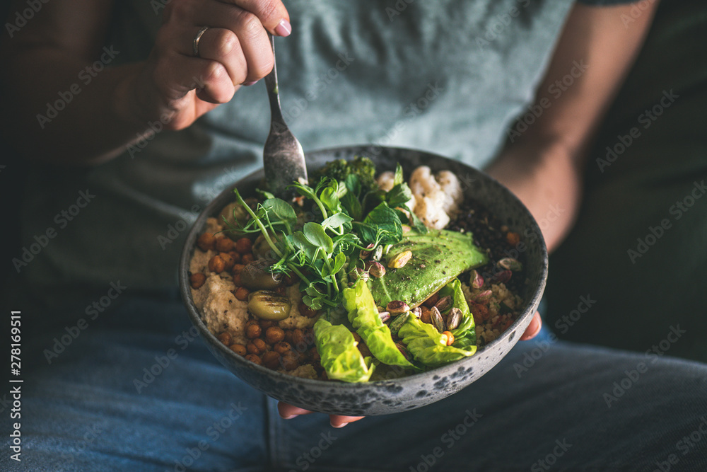 Healthy dinner or lunch. Woman in t-shirt and jeans eating vegan superbowl or Buddha bowl with hummus, vegetable, salad, beans, couscous and avocado - obrazy, fototapety, plakaty 