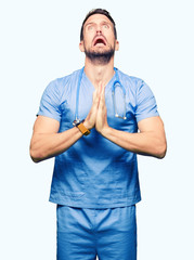 Sticker - Handsome doctor man wearing medical uniform over isolated background begging and praying with hands together with hope expression on face very emotional and worried. Asking for forgiveness. 