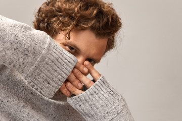 Wall Mural - Attractive hipster guy with wavy ginger hair crossing fingers over his face and looking at camera with penetrating facial expression, dressed in gray pullover. Copyspace for your information