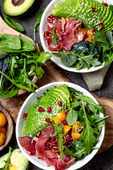 Wall Mural - Low carbs bowl. Fresh salad with green spinach, rucola, avocado an ham serrano in white bowl, gray background, top view