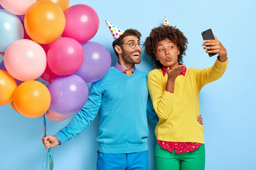 Smiling beautiful Afro girl and her happy boyfriend holds bunch of colorful air balloons, take selfie on smartphone, woman blows airkiss, wear party hats, enjoy festive occasion, pose indoor