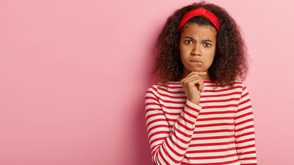 Pensive African American woman holds chin, bites lower lip, has serious expression, looks with worried expression, wears white and red striped jumper, poses against pink wall with blank empty space