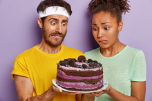 Puzzled Husband And Wife Stare With Big Appetite At High Energy Cake, Bite Lips, Have Desire To Eat, Cannot Afford Dessert As Lead Healthy Lifestyle And Nutrition. People, Junk Food, Temptation
