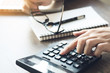 © satapatms - Business accountant man is using the calculator to calculate the income while holding the eye glasses because of stress. Calculate the financial cost concept.