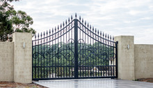 Black Metal Wrought Iron Driveway Property Entrance Gates Set In Concrete Brick Fence, Lights, Garden Trees In Background