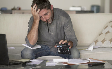 stressed and desperate man at home living room couch doing domestic accounting with paperwork and calculator feeling overwhelmed and worried suffering financial crisis