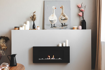 Flowers in vases, candles and rustic painting on shelf above eco fireplace in fashionable living room interior