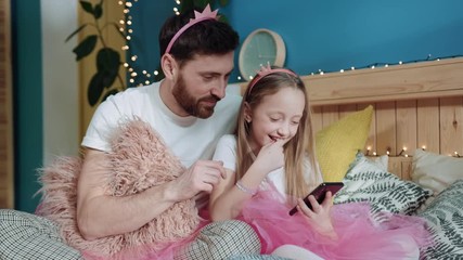 Canvas Print - Beautiful family portrait of cheerful daddy and his little girl using a smartphone while sitting on bed. Father and daughter dressed like magic fairies playing together at home. Family party