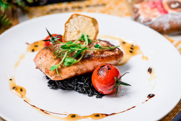 Beautiful serving of roasted salmon fillet and risotto with cuttlefish ink on a white plate with a signature sauce and Cherry tomatoes in a restaurant. close up