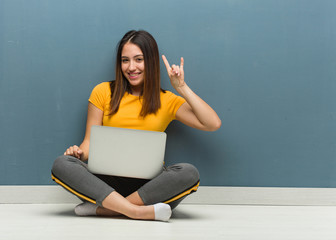 Young woman sitting on the floor with a laptop doing a rock gesture