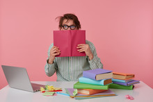 Studio Shot Of Young Man Guy With Glasses, Wears On Blank Shirt, Sitting At A Table With Books, Working At A Laptop, Peeps In Surprise At The Book That Covers Hes Face, Isolated Over Pink Background.