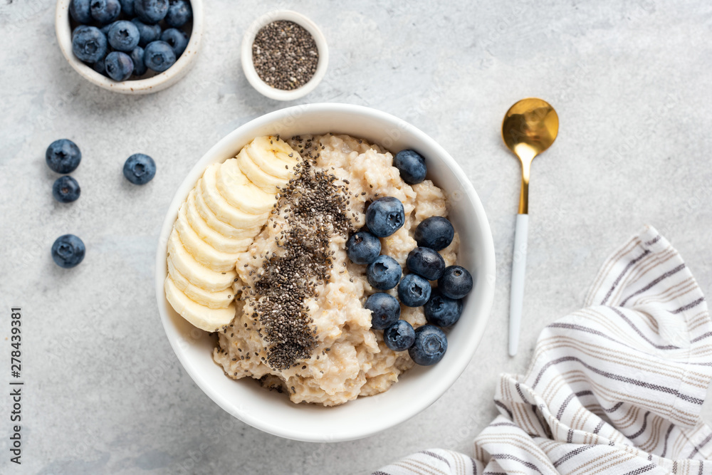 Breakfast oatmeal porridge with banana, blueberries, chia seeds on grey concrete background. Table top view of healthy food for weight loss, clean eating, vegan and vegetarian diet - obrazy, fototapety, plakaty 
